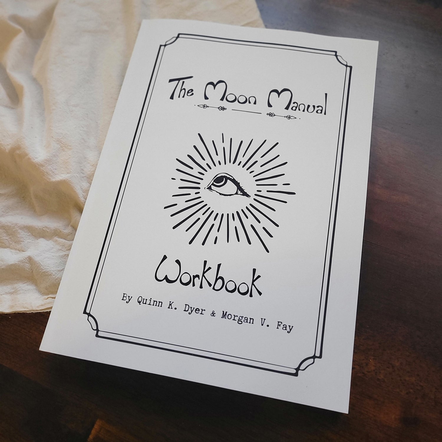A white workbook titled 'The Moon Manual Workbook' with a black hand-drawn eye and sunburst illustration, photographed on a dark wooden surface with a cream fabric cloth underneath.