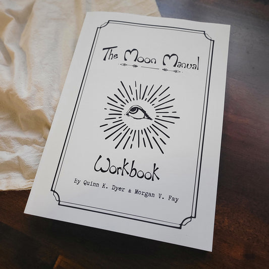A white workbook titled 'The Moon Manual Workbook' with a black hand-drawn eye and sunburst illustration, photographed on a dark wooden surface with a cream fabric cloth underneath.