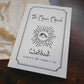A white workbook titled 'The Moon Manual Workbook' with a black hand-drawn eye and sunburst illustration, photographed on a dark wooden surface with a cream fabric cloth underneath.